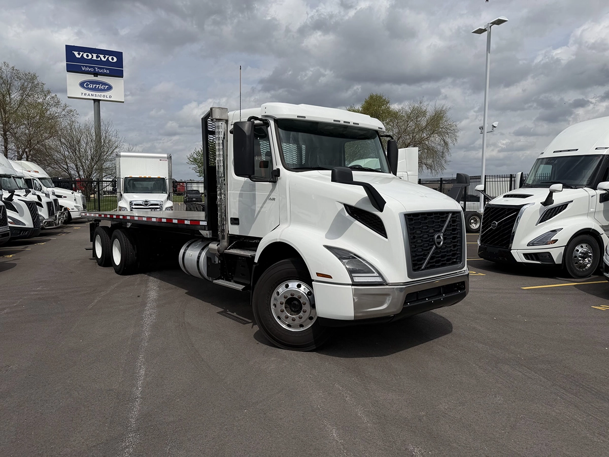 Front driver-side view of a white Volvo VNR flatbed cab and chassis truck with a black flatbed body, parked near Volvo dealership signage.