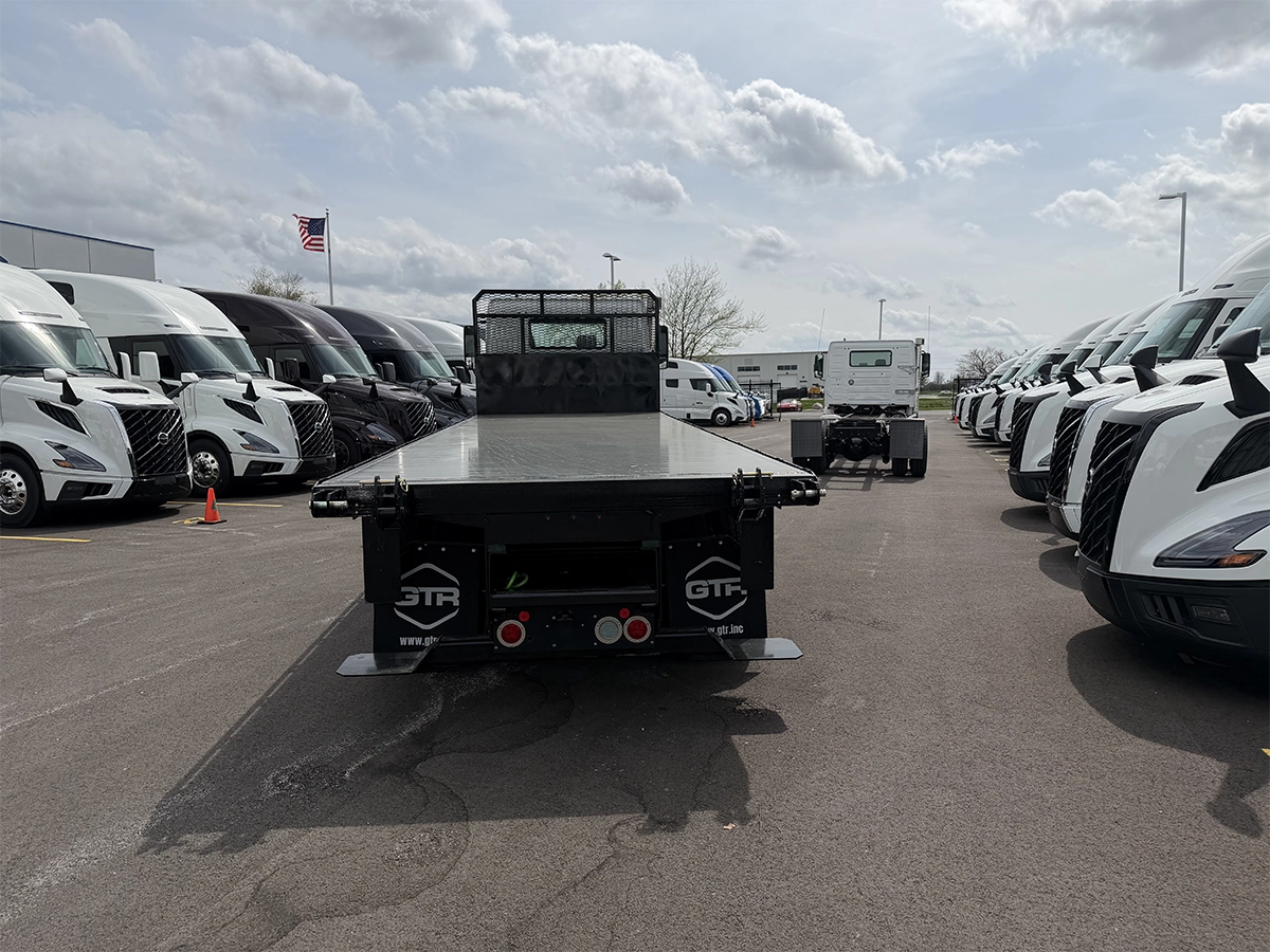 Rear view of a black flatbed truck body with GTR branding and mesh headboard, mounted on a cab and chassis and parked among rows of Volvo sleeper trucks.