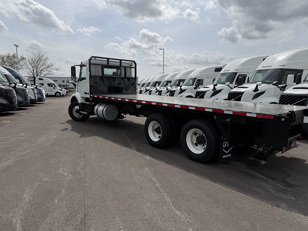 Rear driver-side view of a white flatbed truck with a black bed featuring red and white reflective tape, parked near a row of Volvo sleeper trucks.