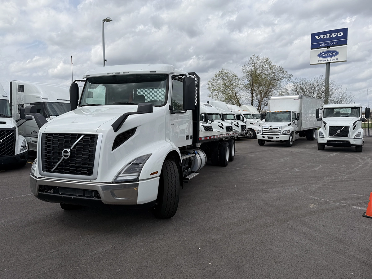 Front passenger-side view of a white Volvo VNR flatbed truck parked at a dealership lot, with a Hino box truck and Volvo/Carrier Transicold sign in the background.