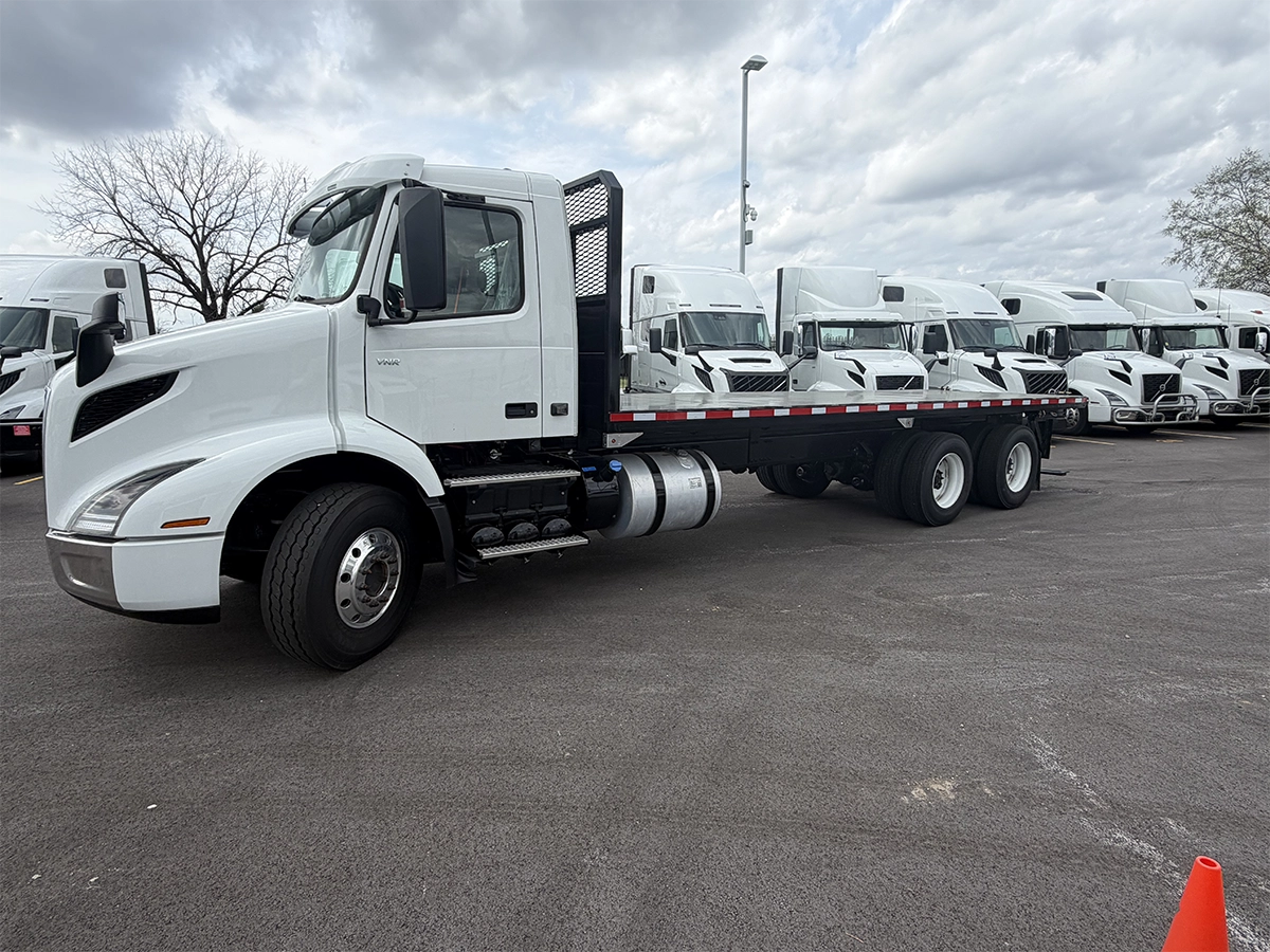 Full passenger-side view of a white Volvo VNR flatbed cab and chassis truck with a black flatbed body, parked alongside rows of Volvo sleeper trucks.