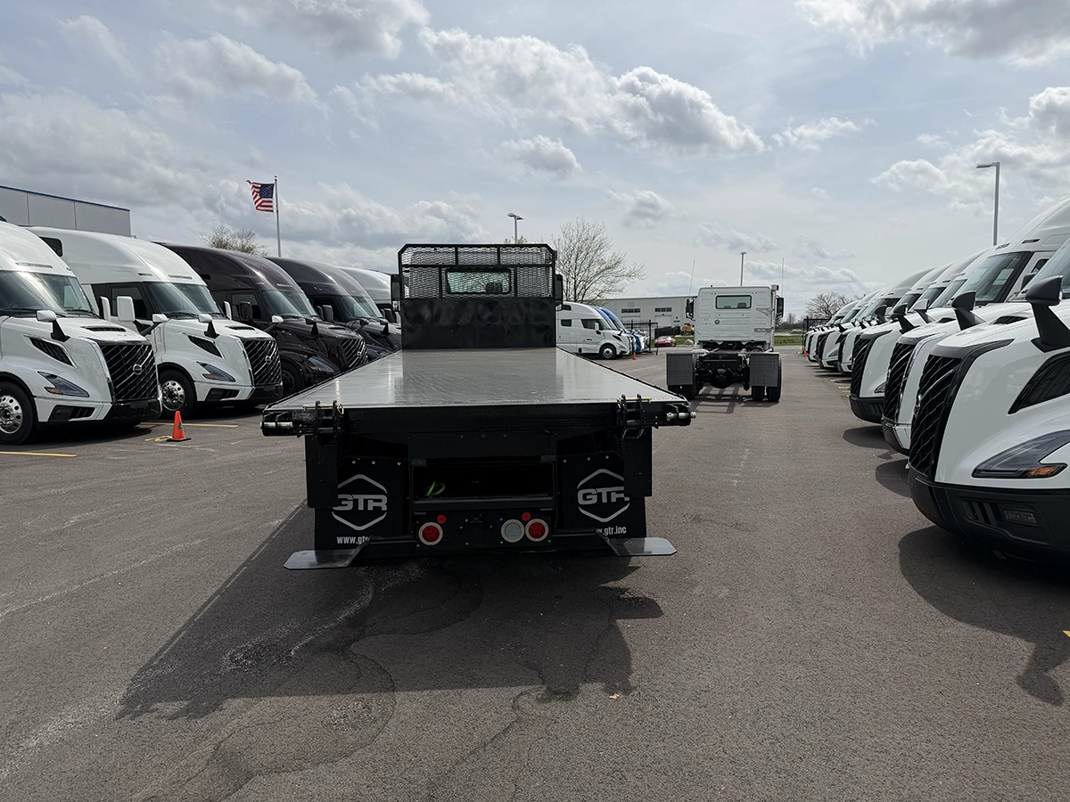 Rear view of a black flatbed truck body with a mesh headache rack, GTR-branded corner plates, and tandem rear axles, parked among other trucks on the Gateway Truck & Refrigeration lot