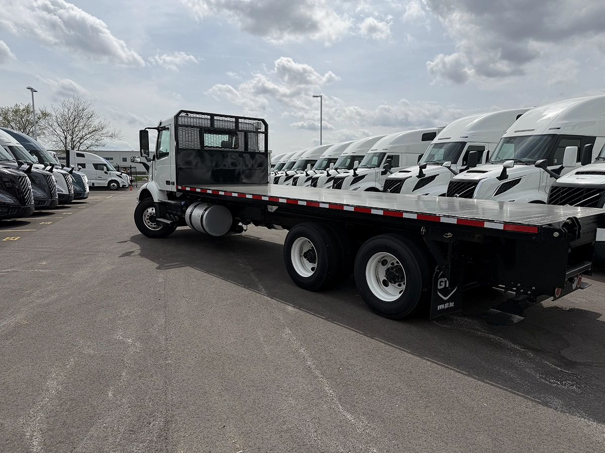 Driver-side profile view of a white flatbed truck with a mesh headache rack, tandem rear axles, and red/white reflective striping, parked at Gateway Truck & Refrigeration.