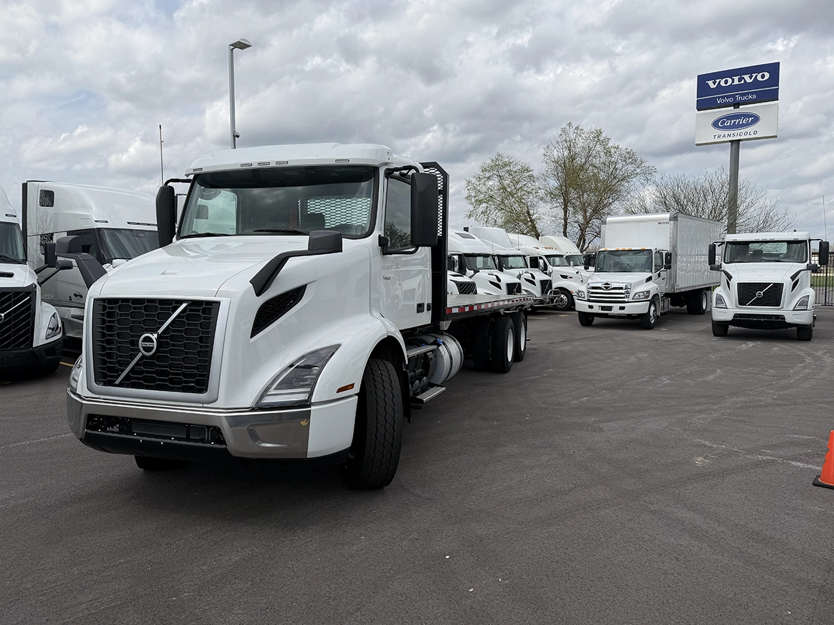 Front driver-side view of a white Volvo flatbed truck with a mesh headache rack, parked at Gateway Truck & Refrigeration with Volvo Trucks and Carrier Transicold signage in the background.