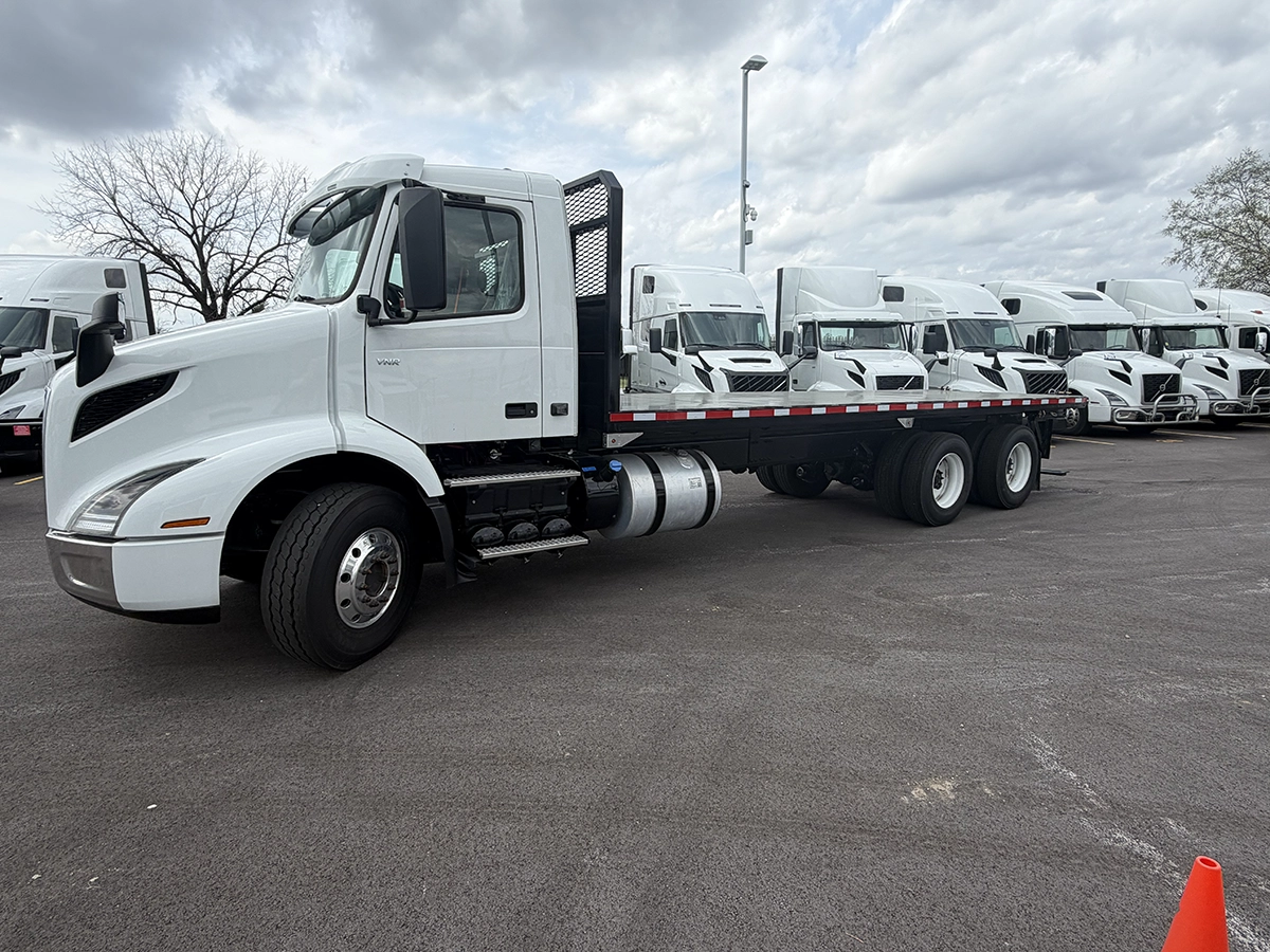 White flatbed truck with a black flatbed trailer parked in a lot, with a row of similar white semi-truck cabs lined up in the background.