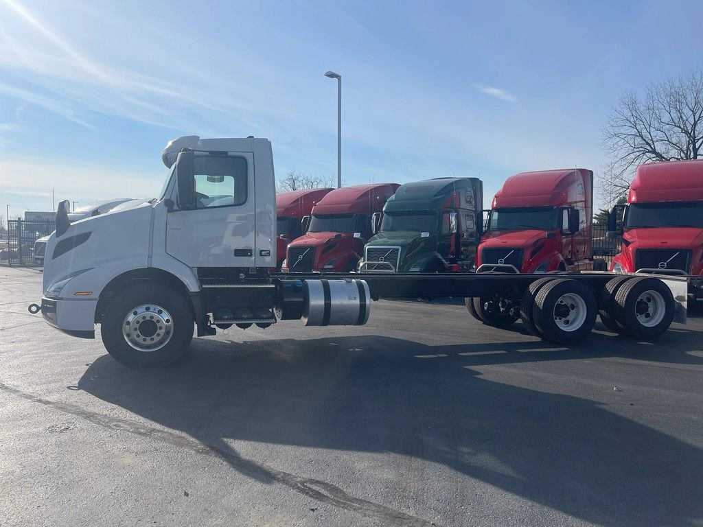 Driver-side view of a white Volvo VNR day cab chassis parked among other trucks on the lot.