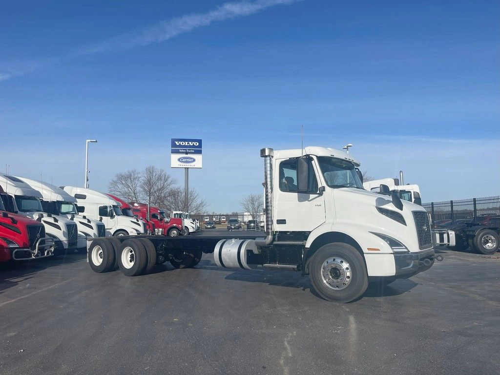 Passenger-side profile of a white Volvo VNR day cab chassis parked among other trucks.