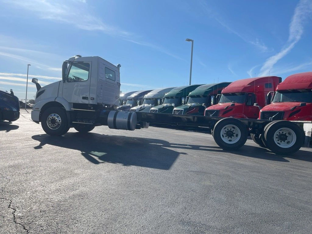 Driver-side profile of a white Volvo VNR day cab chassis parked beside red and white trucks.