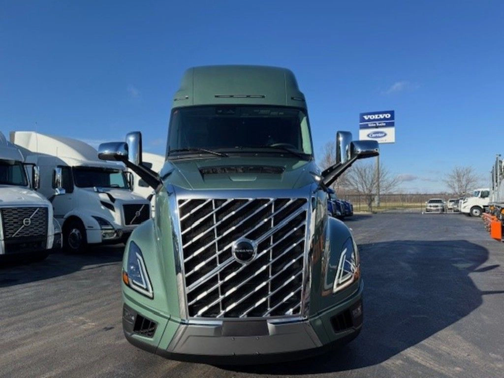 A green Volvo VNL semi-truck is parked on a lot, facing forward, with other trucks and a Volvo dealership sign visible in the background.