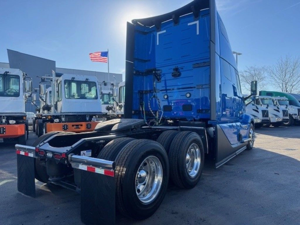 A blue semi-truck cab with gleaming silver wheels is parked in a lot, flanked by white trucks. An American flag waves in the clear blue sky.