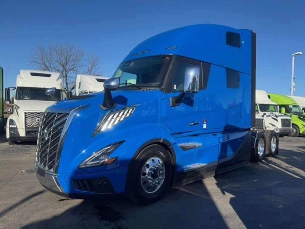 Bright blue semi-truck parked in a lot with a clear sky. Other trucks in white and green are visible in the background. Daytime setting.