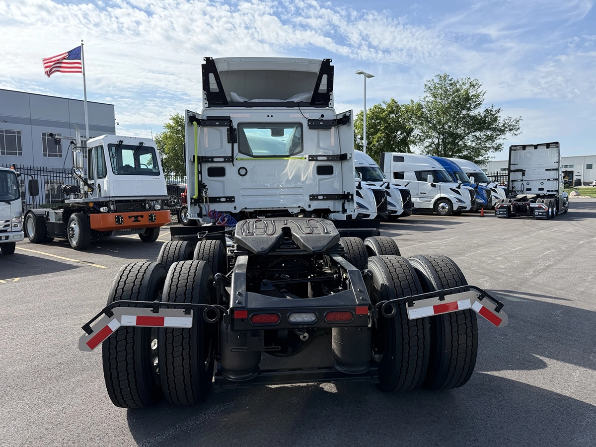 Rear view of a 2027 Volvo VNR64T300 white day cab semi truck, showing the fifth wheel plate, dual rear axles, and tail lights in a dealership lot with an American flag in the background