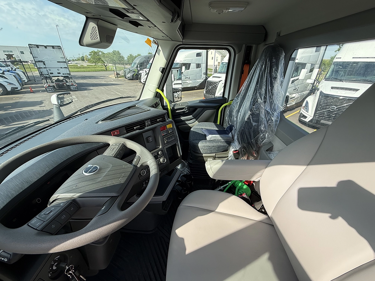 Interior cab view of a 2027 Volvo VNR64T300 showing the driver's seat, Volvo-branded steering wheel with integrated controls, dashboard, and light gray upholstered passenger seat, with the dealership lot visible through the windshield