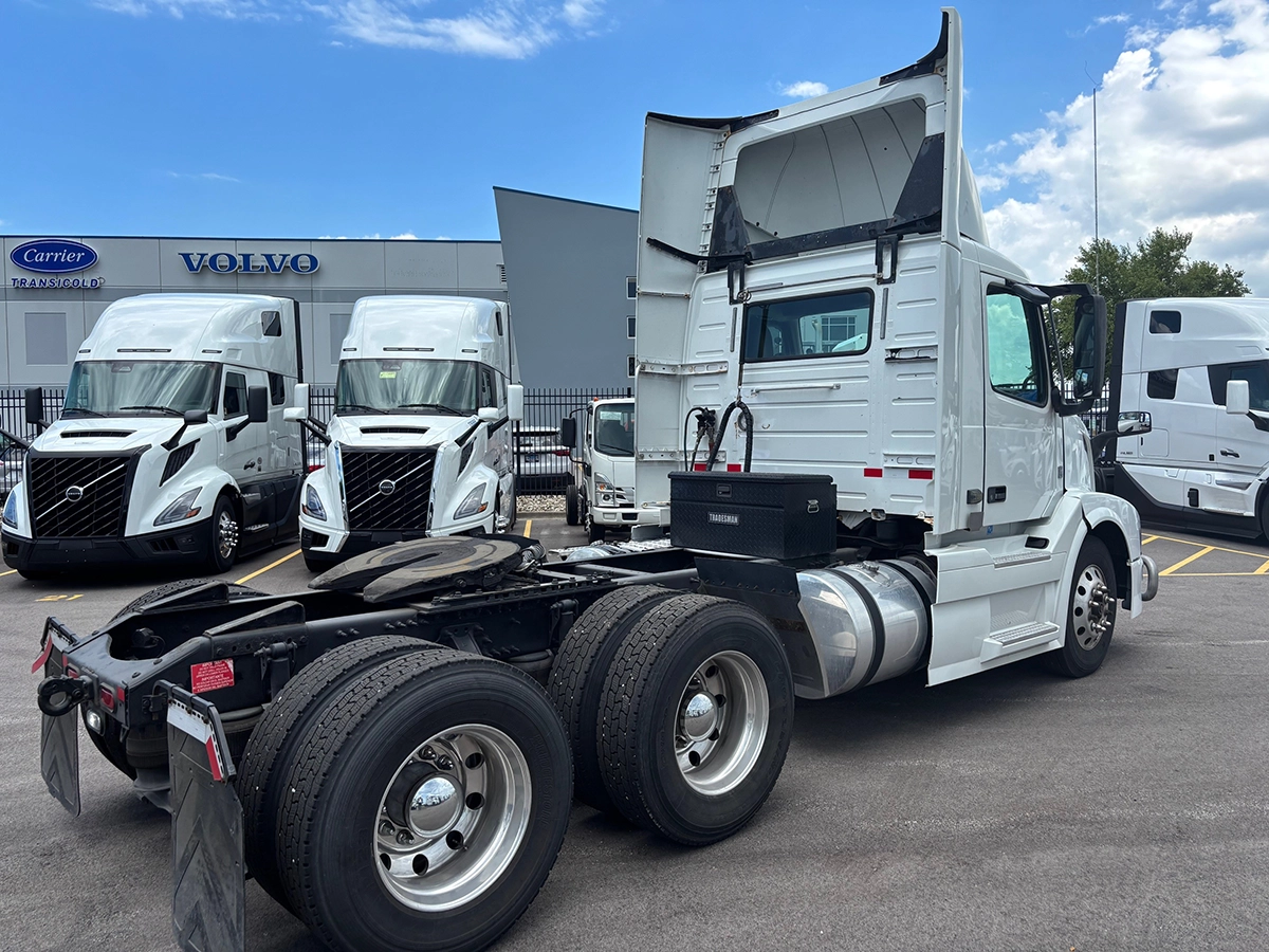 Rear passenger-side view of a white Volvo VNR sleeper cab truck tractor with raised sun visor and toolbox mounted behind the cab, showing tandem axles at the dealership.