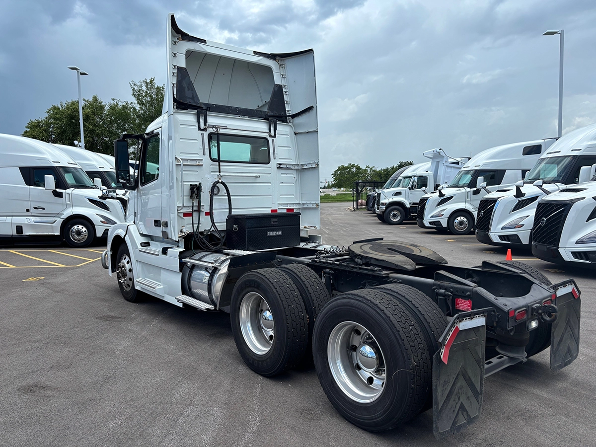 Rear driver-side view of the white Volvo VNR sleeper cab tractor with raised sun visor, showing tandem rear axles and mud flaps, with other trucks parked in the background.