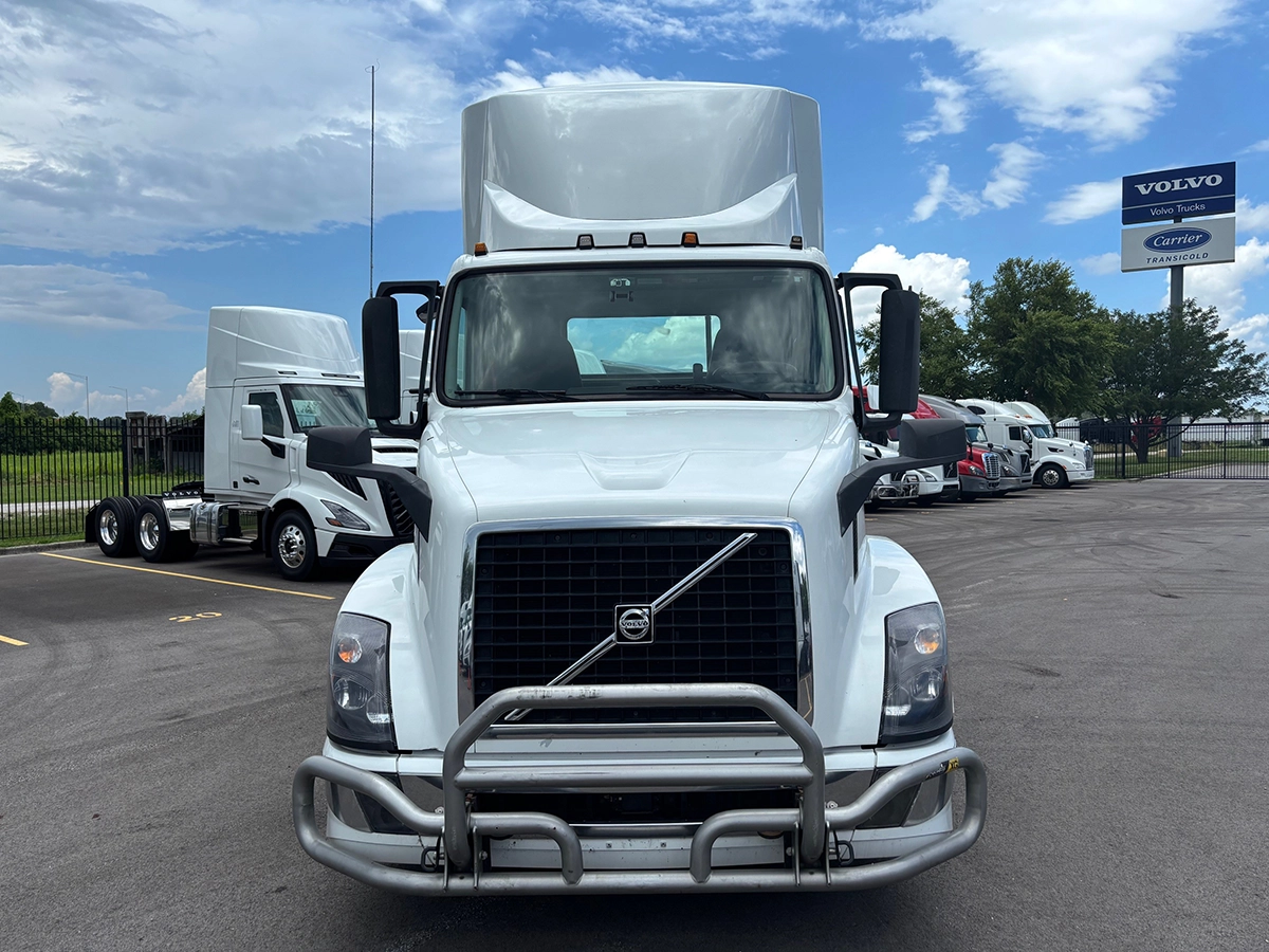 Straight-on front view of the white Volvo VNR sleeper cab truck tractor showing the grille, headlights, and chrome bumper guard, with a Volvo Trucks dealership sign in the background.
