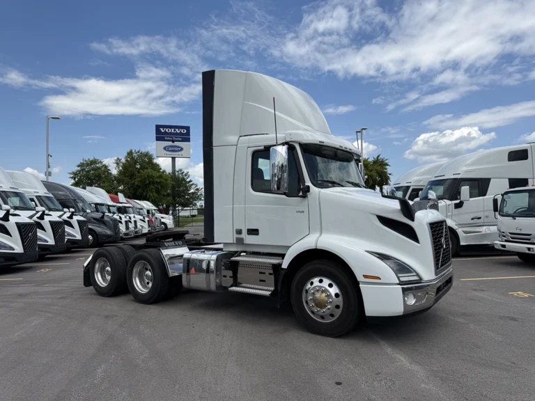 White 2022 Volvo VNR64T300 day cab truck tractor parked at a dealership lot, front three-quarter view, with other trucks and a Volvo Trucks sign visible in the background.