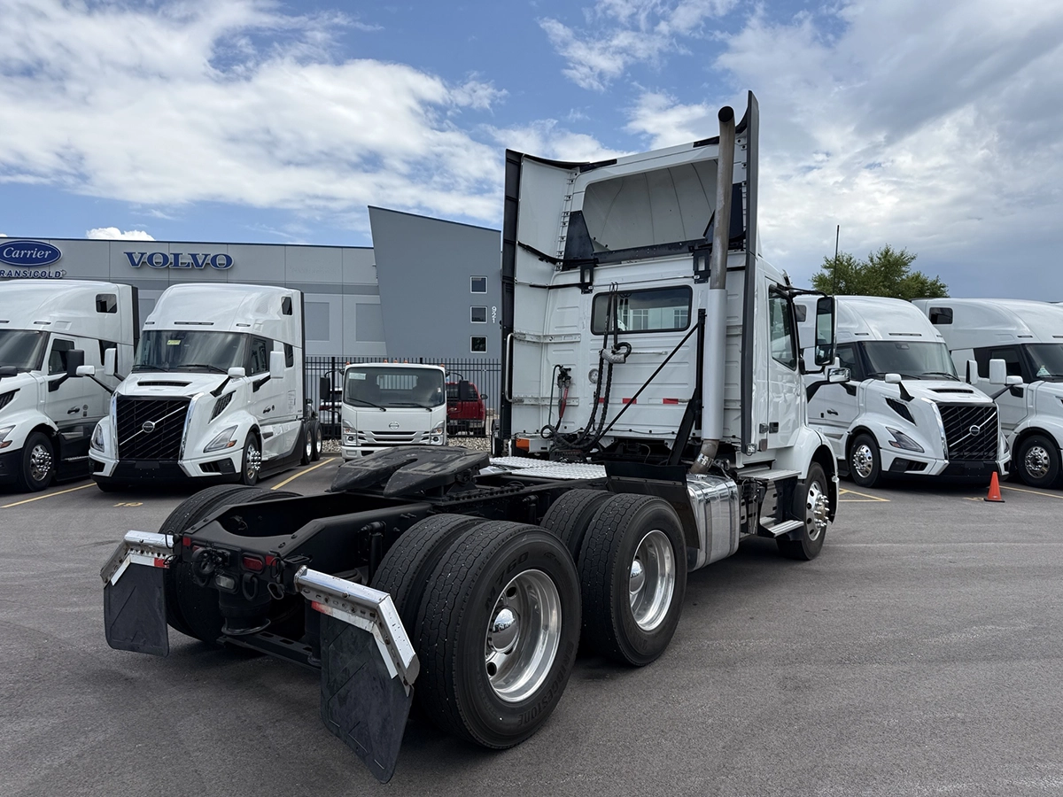 Rear three-quarter view of the white Volvo VNR day cab, showing the open frame rails, tandem axles, and mud flaps, with other trucks parked in the background.