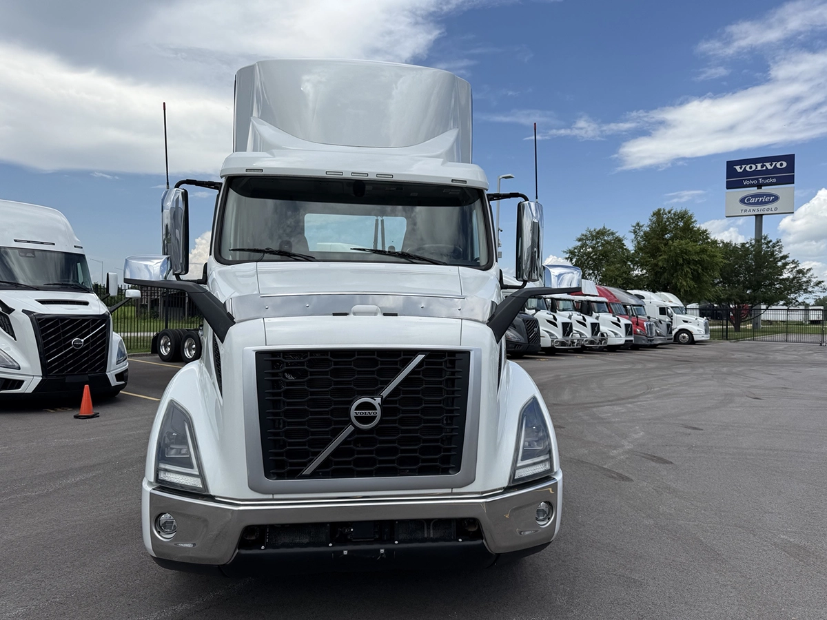Straight-on front view of the white Volvo VNR truck cab showing the grille, headlights, and chrome mirrors, with other trucks and a dealership sign in the background.
