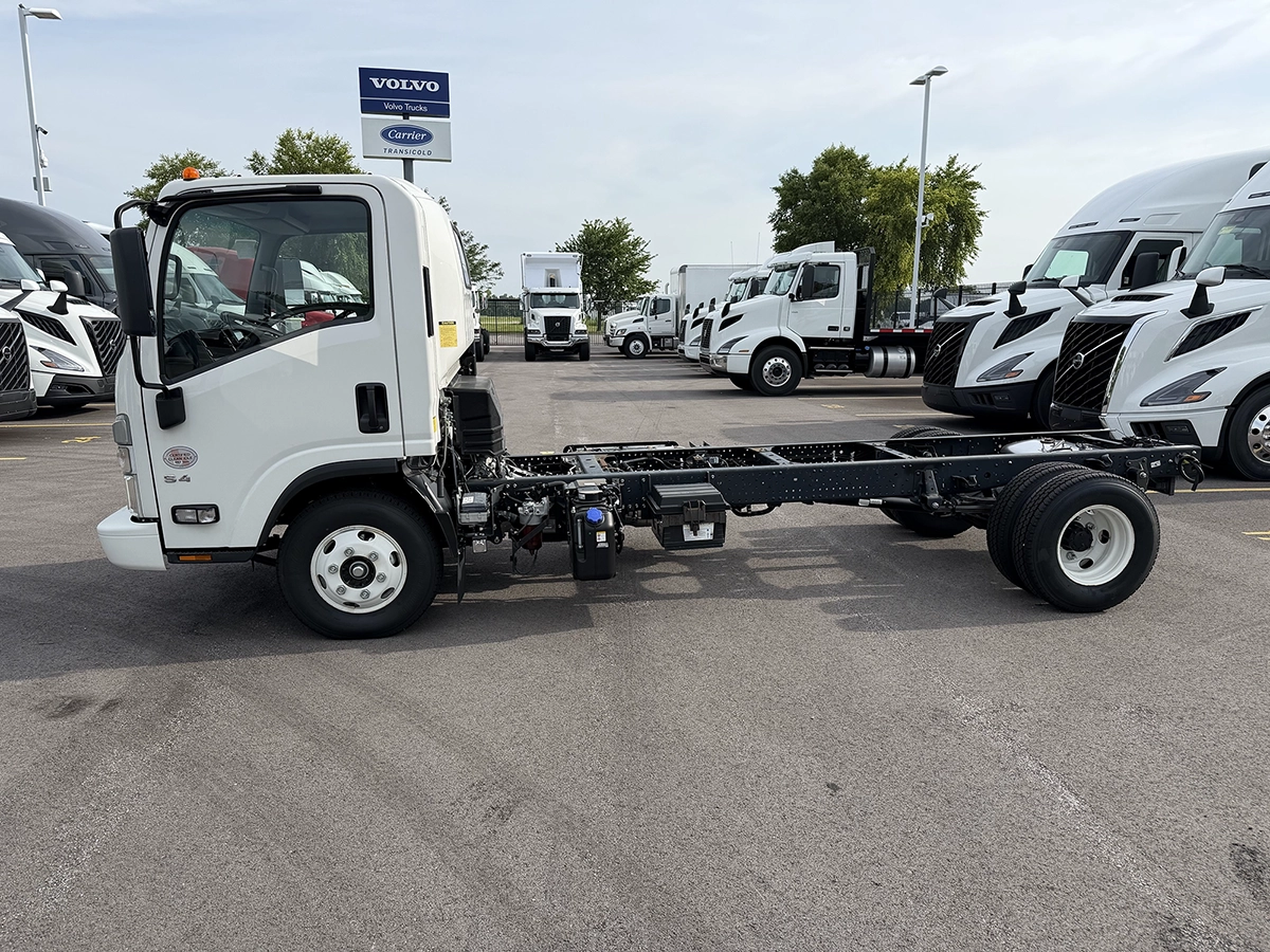 Driver side view of a white 2023 Hino S4 cab and chassis truck with exposed frame rails, parked near Volvo dealership signage.