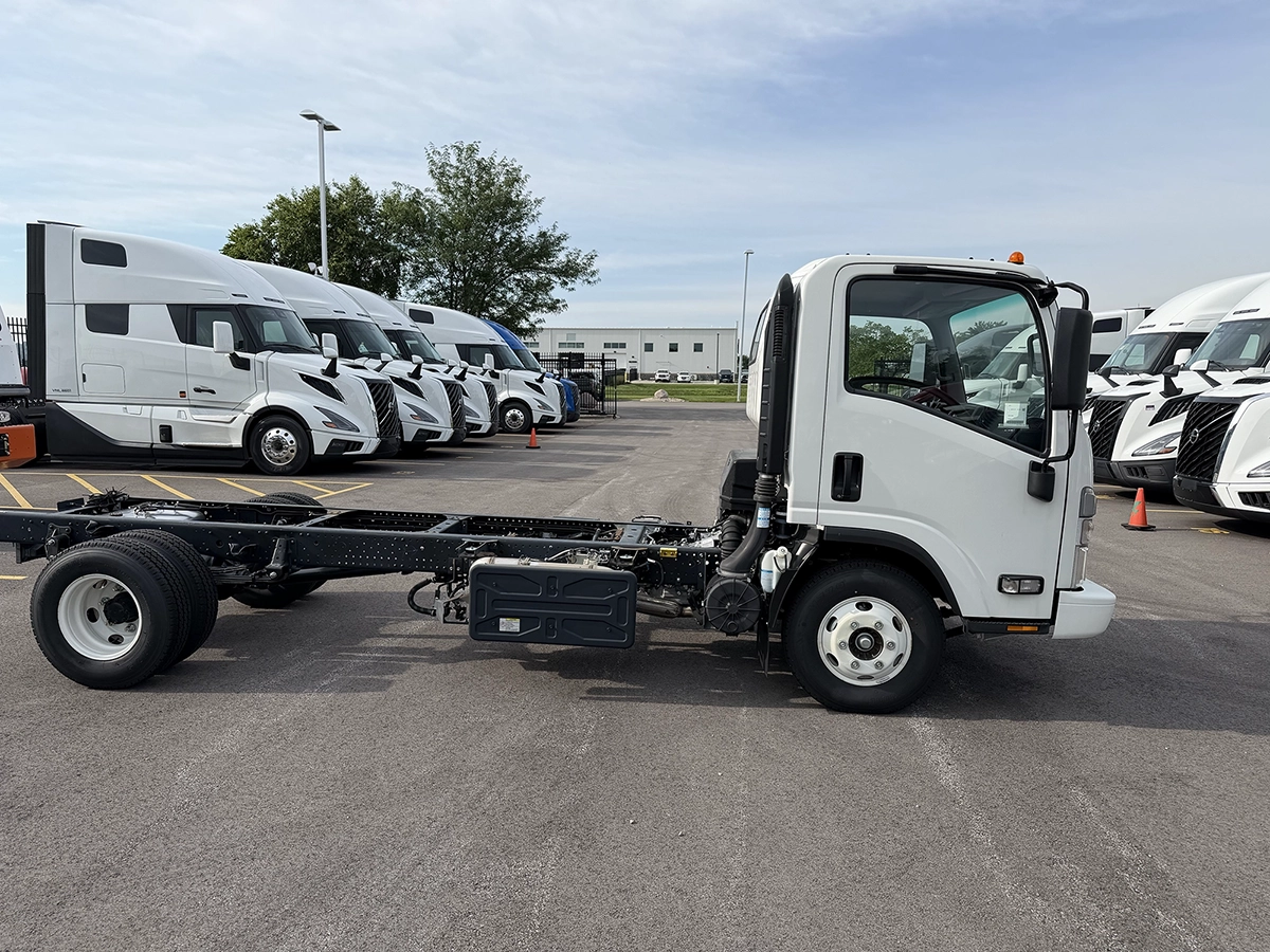 Passenger side view of a white 2023 Hino S4 cab and chassis truck with exposed frame rails, parked near rows of Volvo sleeper trucks.