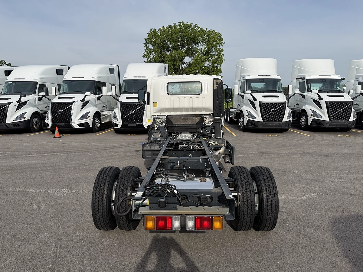 Rear view of a white 2023 Hino S4 cab and chassis truck showing the exposed frame rails and dual rear wheels, parked among rows of Volvo sleeper trucks.