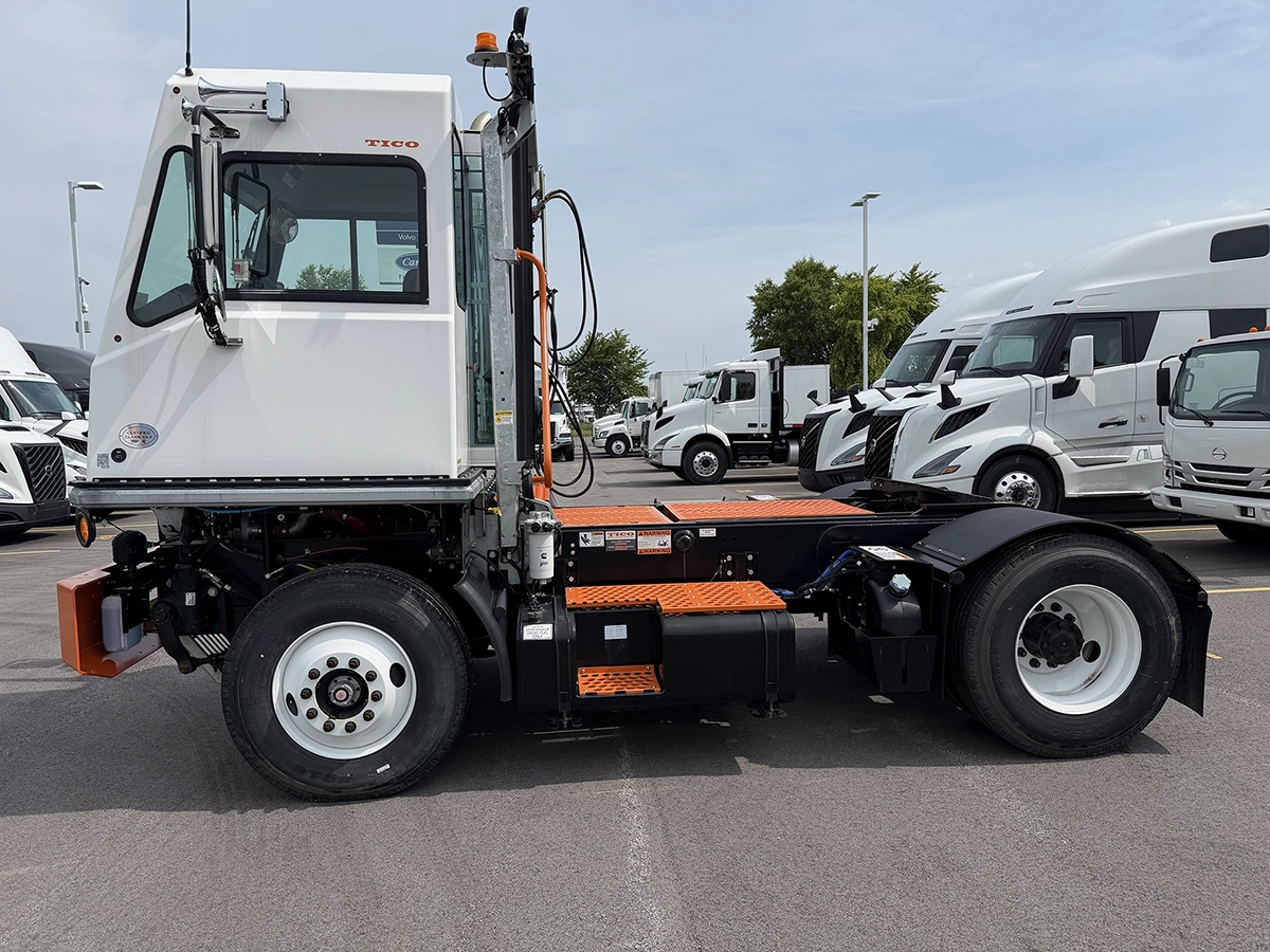 Driver side view of a white 2024 TICO Pro-Spotter yard spotter truck with a raised cab and orange step platforms, parked at a dealership lot.