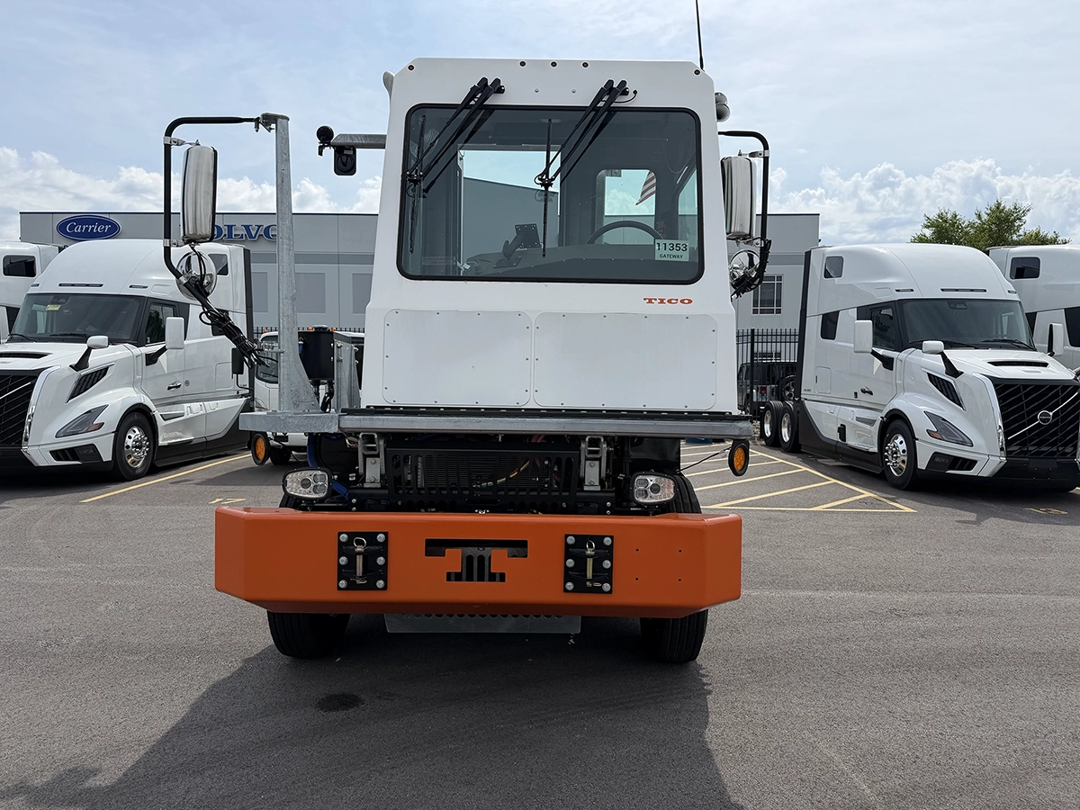 Front view of a white 2024 TICO Pro-Spotter yard truck with an orange bumper and raised cab, parked near Volvo sleeper trucks and dealership signage.