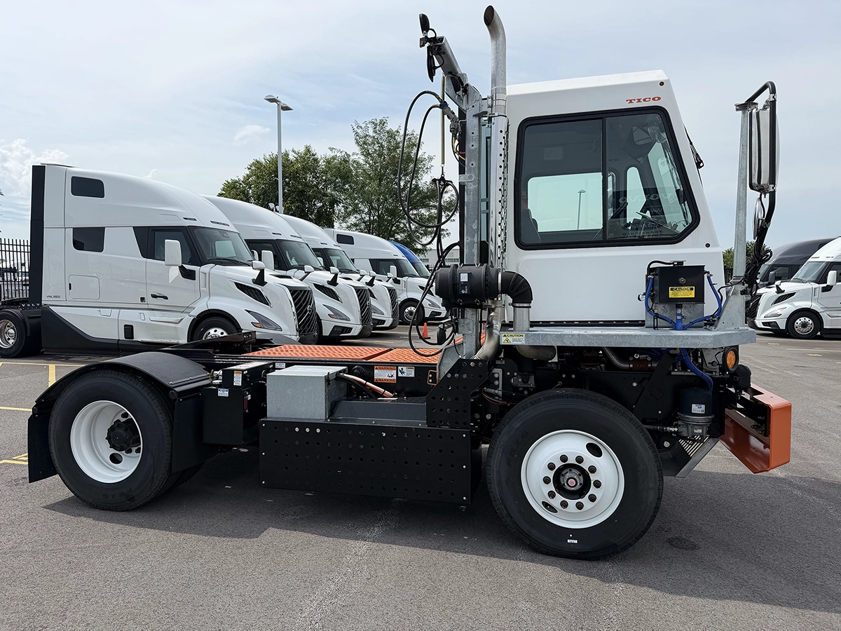 Passenger side view of a white 2024 TICO Pro-Spotter yard spotter truck with a raised cab and orange step platforms, parked at a dealership lot.