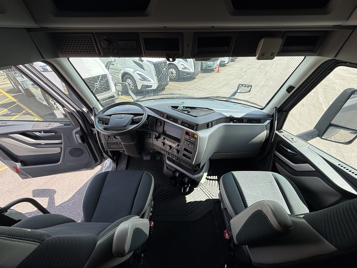 Interior cab view of a 2025 Volvo VNL64T860 showing the steering wheel, digital displays, center dashboard controls, and two cloth seats.