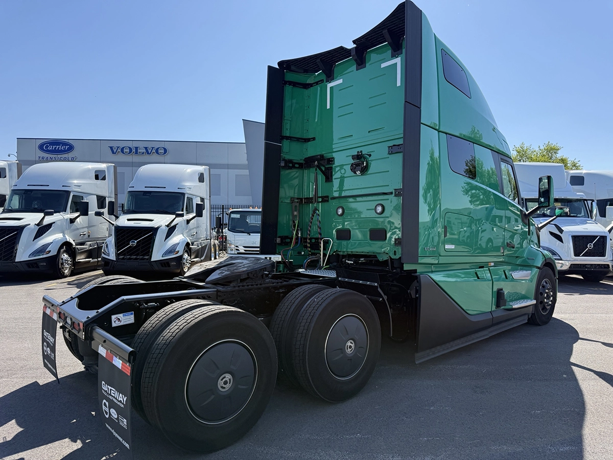 Rear passenger side view of a green 2025 Volvo VNL64T860 showing the fifth wheel, tandem drive axles, and rear of the sleeper cab.