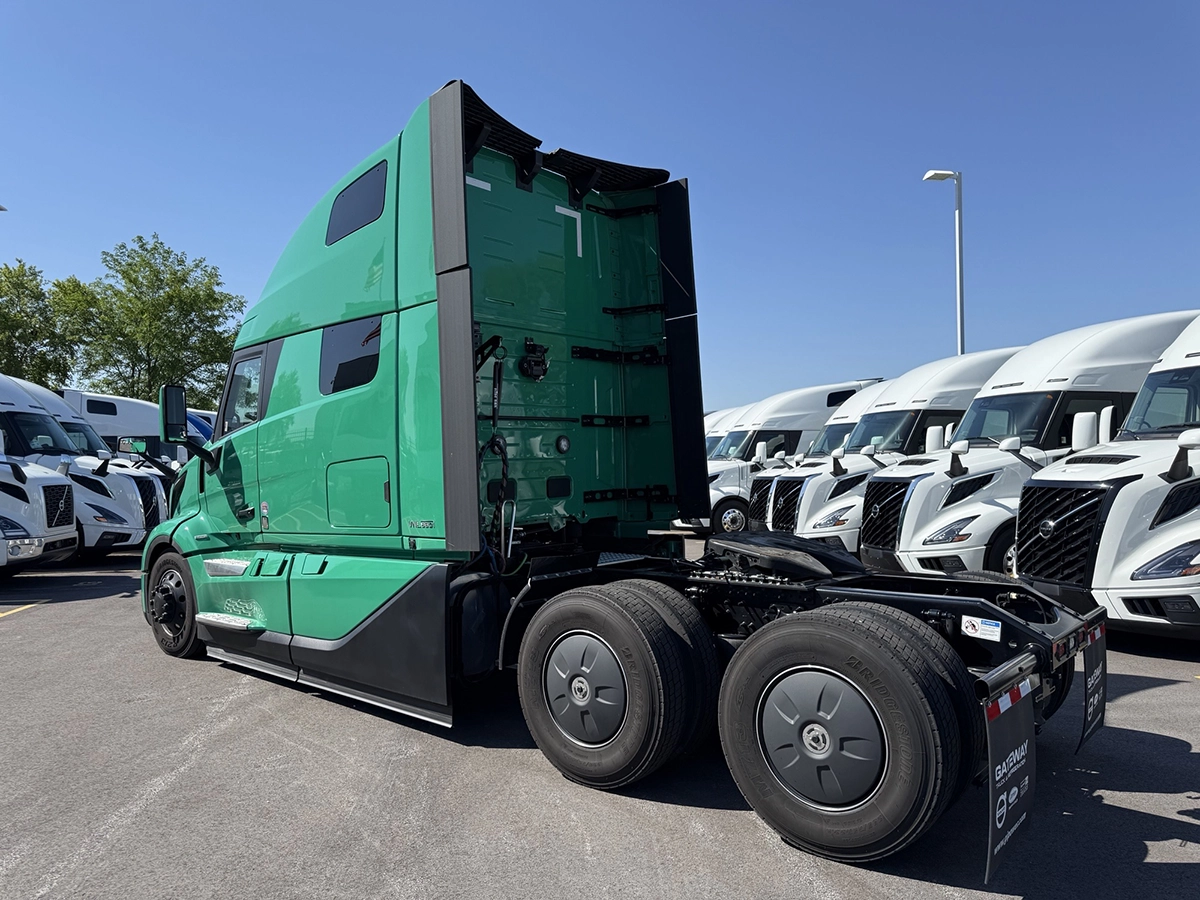 Rear driver side view of a green 2025 Volvo VNL64T860 sleeper semi truck showing the aerodynamic cab extenders and tandem drive axles.