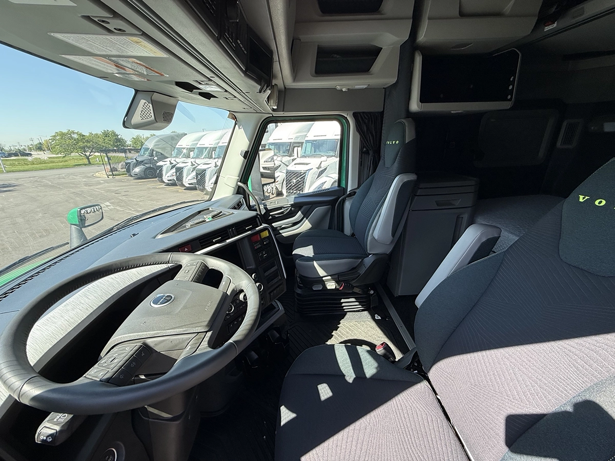 Interior cab view of a 2025 Volvo VNL64T860 showing the steering wheel, gray cloth seats with Volvo embroidery, and sleeper entry.