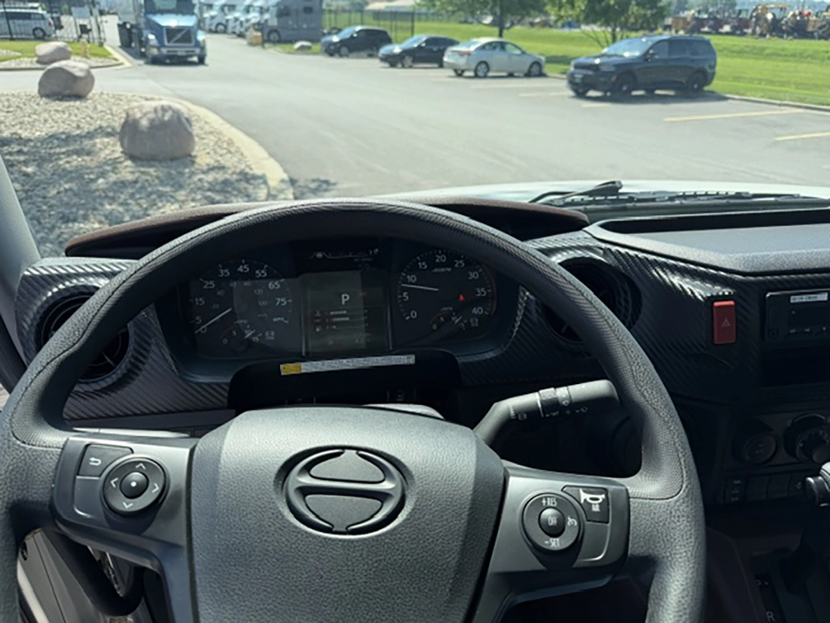 Dashboard view of a 2026 Hino L6 from behind the steering wheel, showing the gauge cluster with digital display, speedometer, and steering wheel controls.