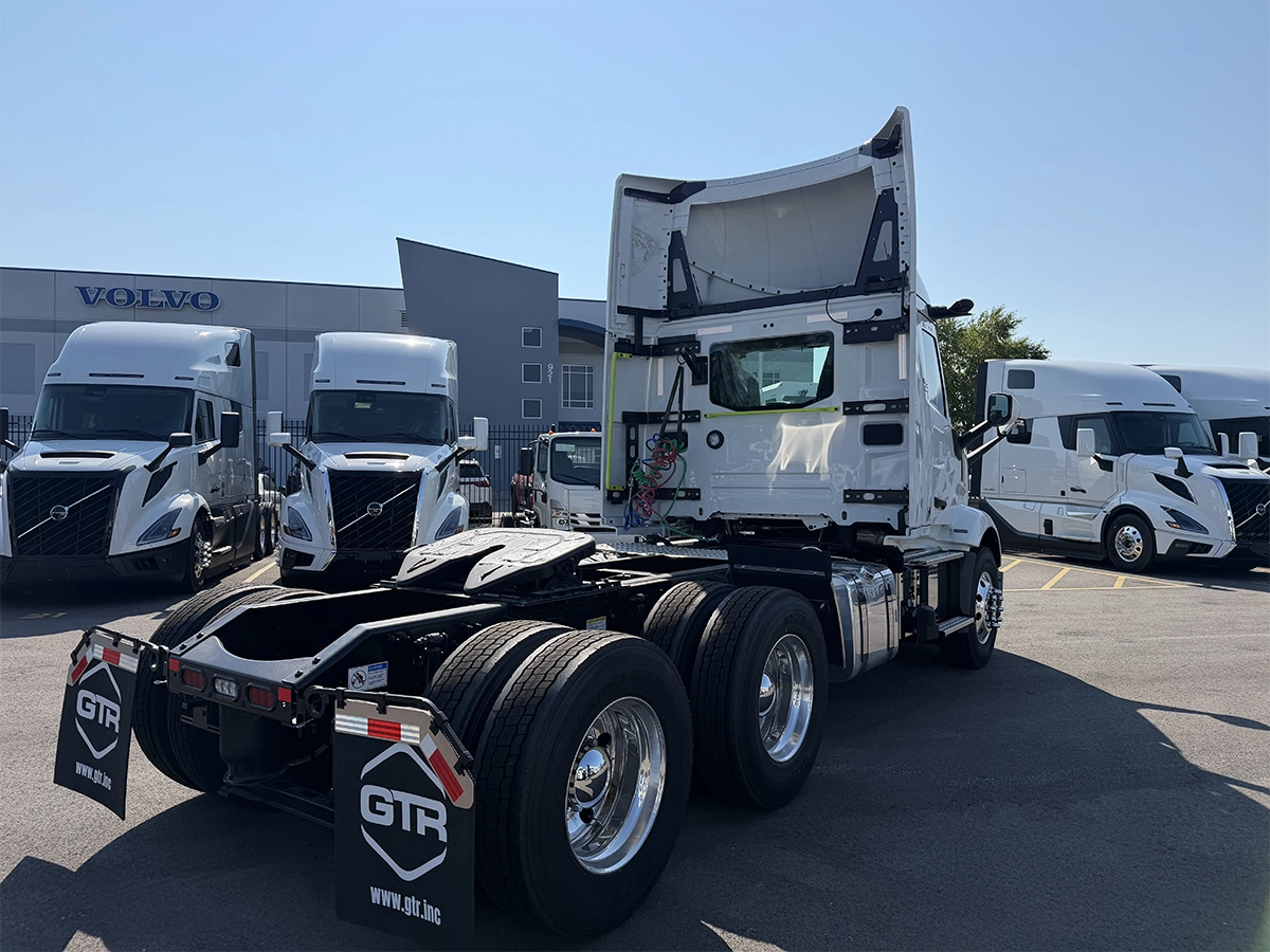 Rear passenger-side view of a white Volvo day cab truck tractor with the cab tilted forward, showing tandem rear axles, chrome wheels, and GTR-branded mud flaps, parked at the Gateway Truck & Refrigeration Collinsville lot with a Volvo dealership sign in the background.